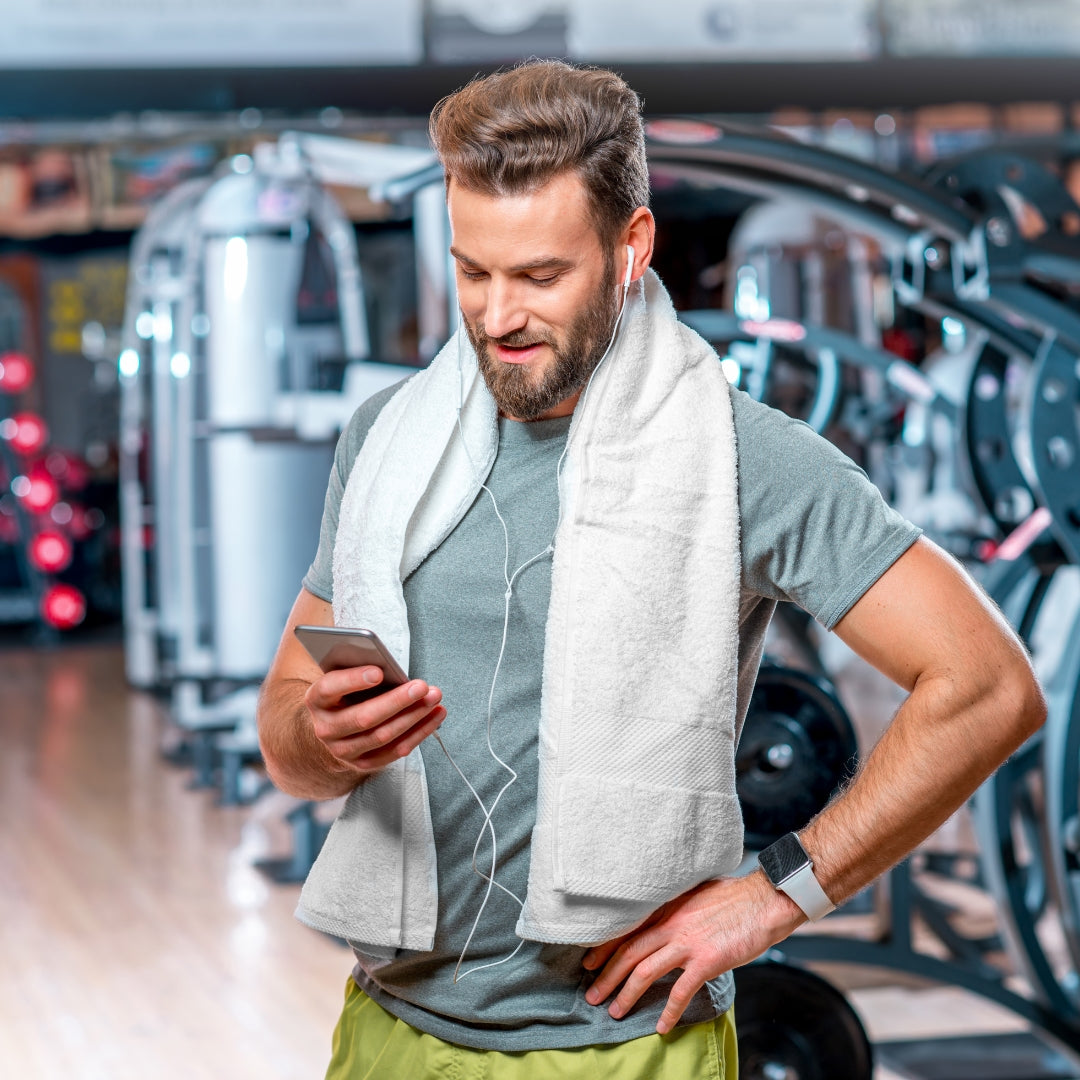 Man with a towel draped over his shoulder using a smartphone in a gym.