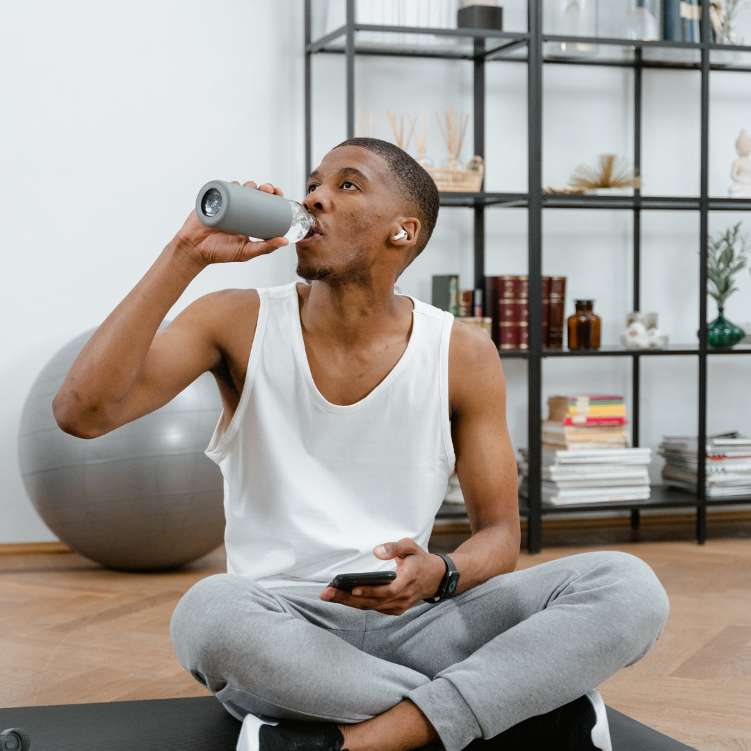 Man sitting on a yoga mat in a home setting, drinking from a water bottle.